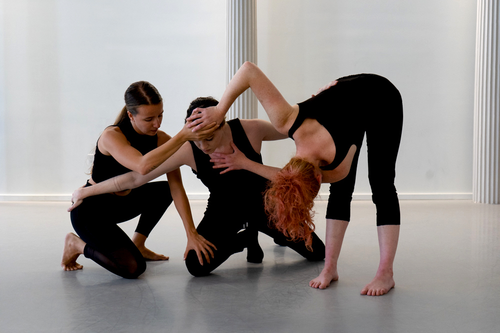 Three dancers kneel and place their hands on each other
